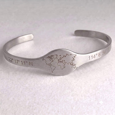 A brushed silver metal cuff bracelet featuring an engraved world map in the center and geographic coordinates on both ends, displayed on a light textured background.