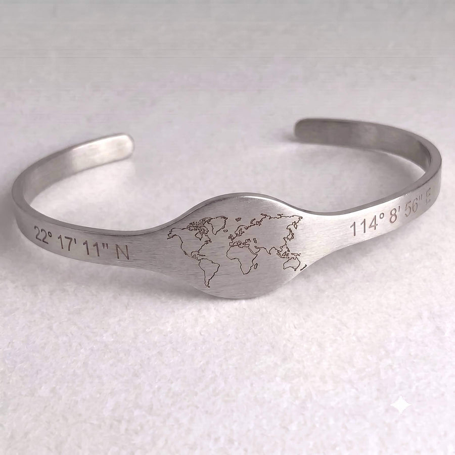 A gold-colored open cuff bracelet featuring an engraved world map in the center, flanked by engraved geographic coordinates, with two small clear rhinestones on a light textured background.
