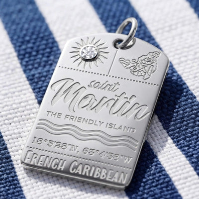 A silver Saint Martin luggage tag pendant with a small clear crystal, engraved with the text 'Saint Martin', 'The Friendly Island', coordinates, and 'French Caribbean', resting on a blue and white striped fabric background.