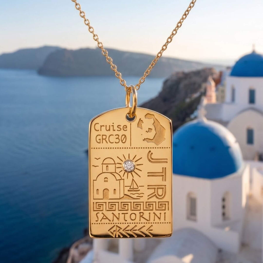 Two travel-themed charms displayed outdoors on a stone ledge with the Acropolis of Athens in the background. The gold charm is a multi-directional road sign pointing to Athens, Corfu, Santorini, and Mykonos. The silver charm is shaped like a postcard with the engraving of the Greek flag and the word Opa.