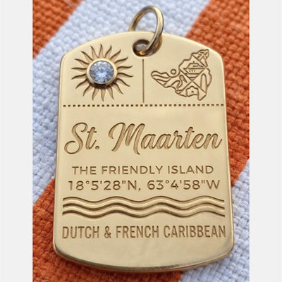 A gold St. Maarten luggage tag charm featuring engraved text, a sun design with a clear rhinestone, and a small map illustration, resting on a textured orange and white striped fabric background.