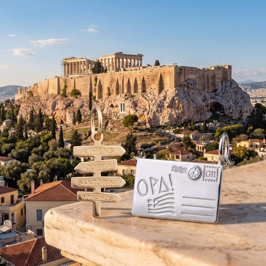 Two travel-themed charms displayed outdoors on a stone ledge with the Acropolis of Athens in the background. The gold charm is a multi-directional road sign pointing to Athens, Corfu, Santorini, and Mykonos. The silver charm is shaped like a postcard with the engraving of the Greek flag and the word Opa.