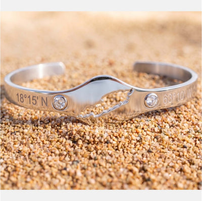 A silver cuff bracelet with a cut-out map of Anguilla featuring two clear gemstones and engraved coordinates '18°15' N' and '63°10' W' on a sandy beach background.