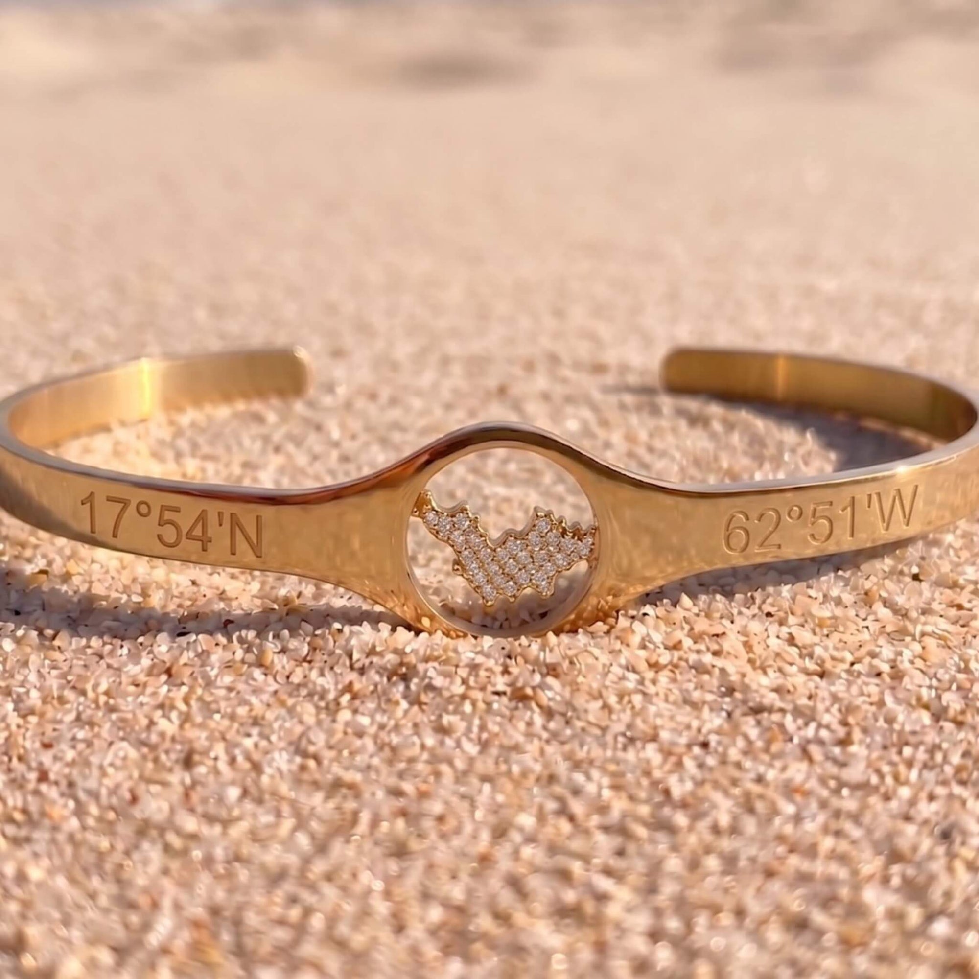 A gold-tone cuff bracelet engraved with geographic coordinates '17°54'N' and '62°51'W', featuring a central oval cutout filled with a pavé diamond map of St-Barth, resting on sandy beach.