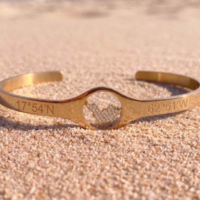 A gold-tone cuff bracelet engraved with geographic coordinates '17°54'N' and '62°51'W', featuring a central oval cutout filled with a pavé diamond map of St-Barth, resting on sandy beach.
