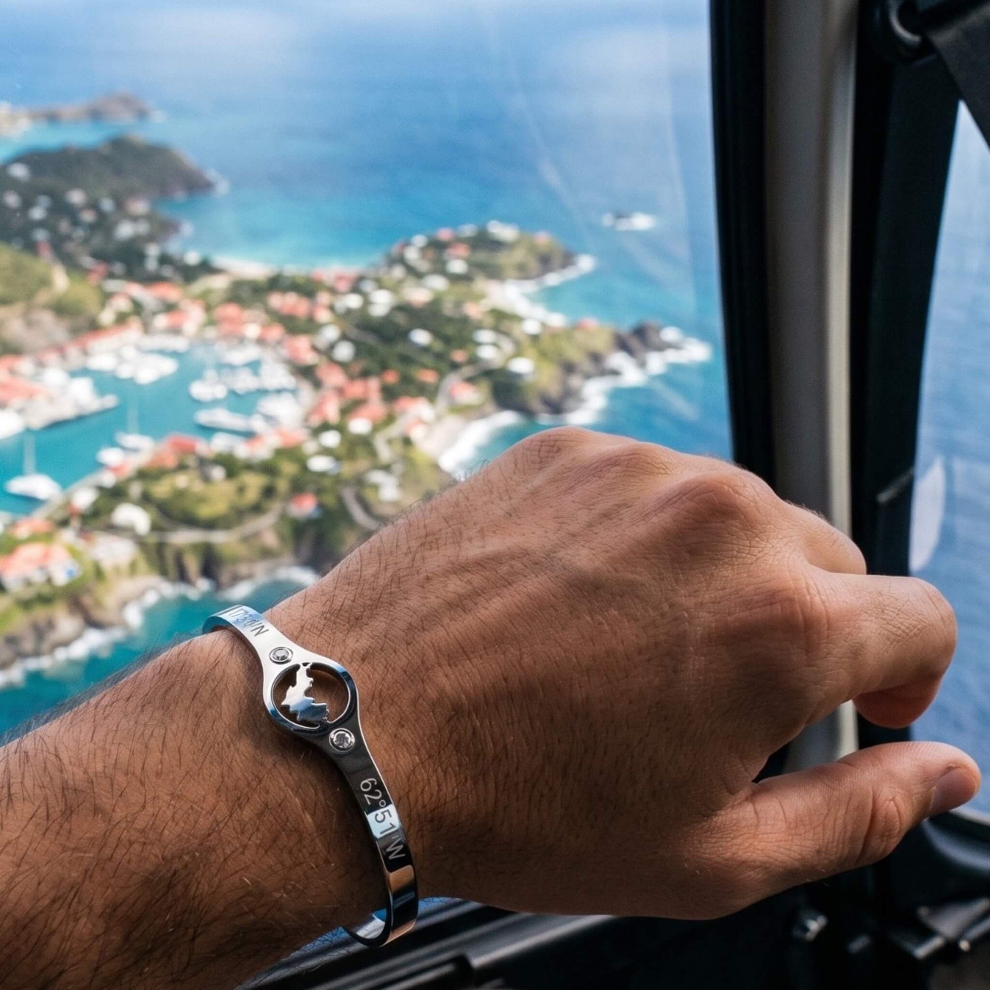 A silver cuff bracelet engraved with coordinates and featuring the map of St-Barth, worn on a man's wrist on board an helicopter over the island of St-Barth.