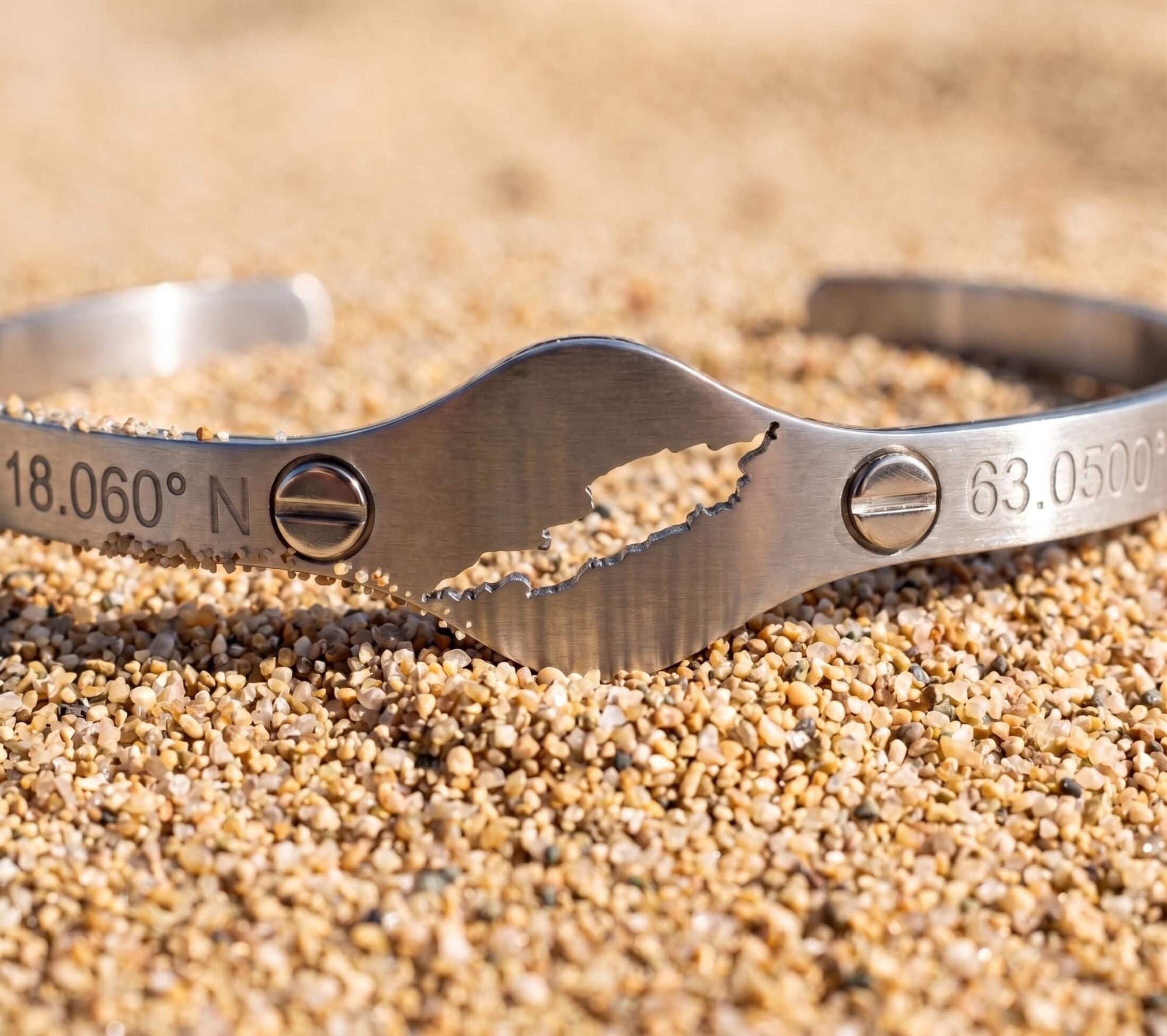 A brushed steel cuff bracelet with a cut-out map of Anguilla featuring two clear gemstones and engraved coordinates '18°15' N' and '63°10' W' on a sandy beach background.