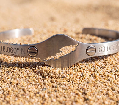 A brushed steel cuff bracelet with a cut-out map of Anguilla featuring two clear gemstones and engraved coordinates '18°15' N' and '63°10' W' on a sandy beach background.