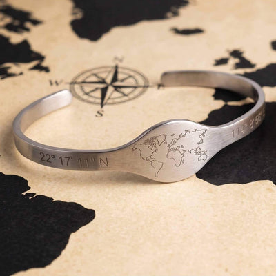 A brushed silver cuff bracelet engraved with a world map and coordinates, displayed on a vintage-style world map background.