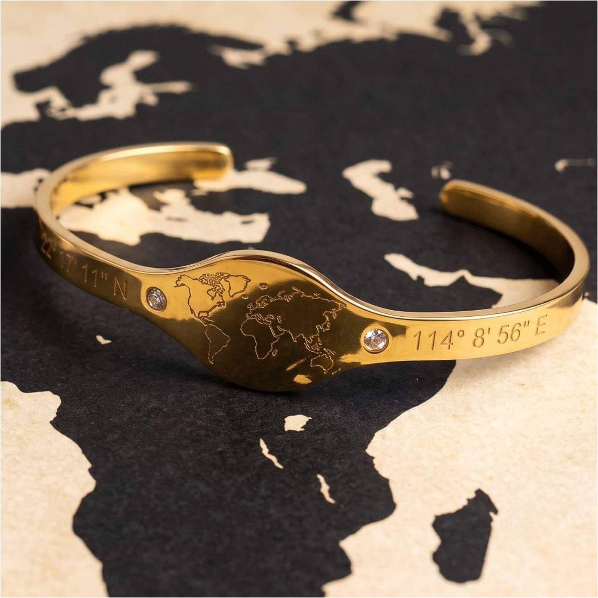 A gold cuff bracelet featuring an engraved world map in the center, flanked by coordinates and accented with two small clear rhinestones, displayed on a black and beige world map background.