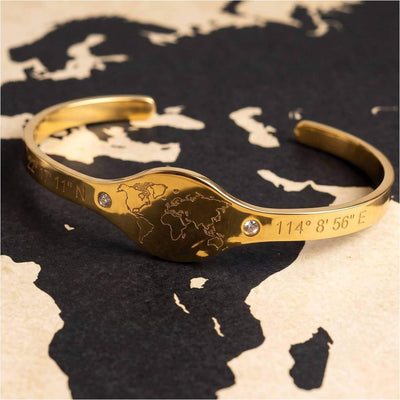A gold cuff bracelet featuring an engraved world map in the center, flanked by coordinates and accented with two small clear rhinestones, displayed on a black and beige world map background.