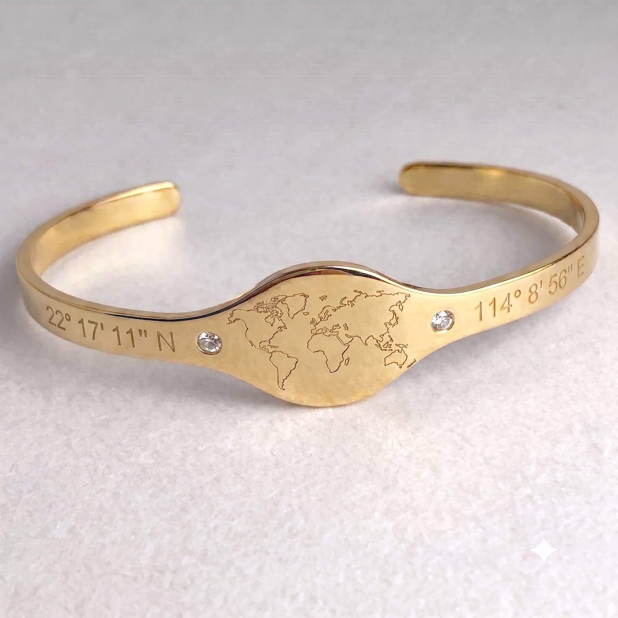 A gold-colored open cuff bracelet featuring an engraved world map in the center, flanked by engraved geographic coordinates, with two small clear rhinestones on a light textured background.