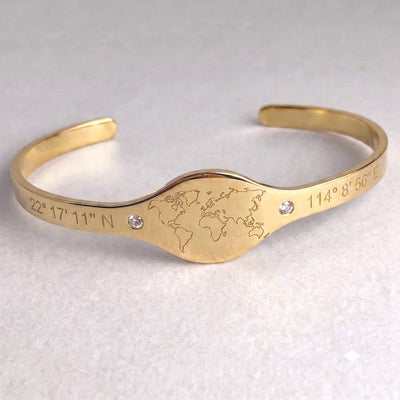 A gold-colored open cuff bracelet featuring an engraved world map in the center, flanked by engraved geographic coordinates, with two small clear rhinestones on a light textured background.
