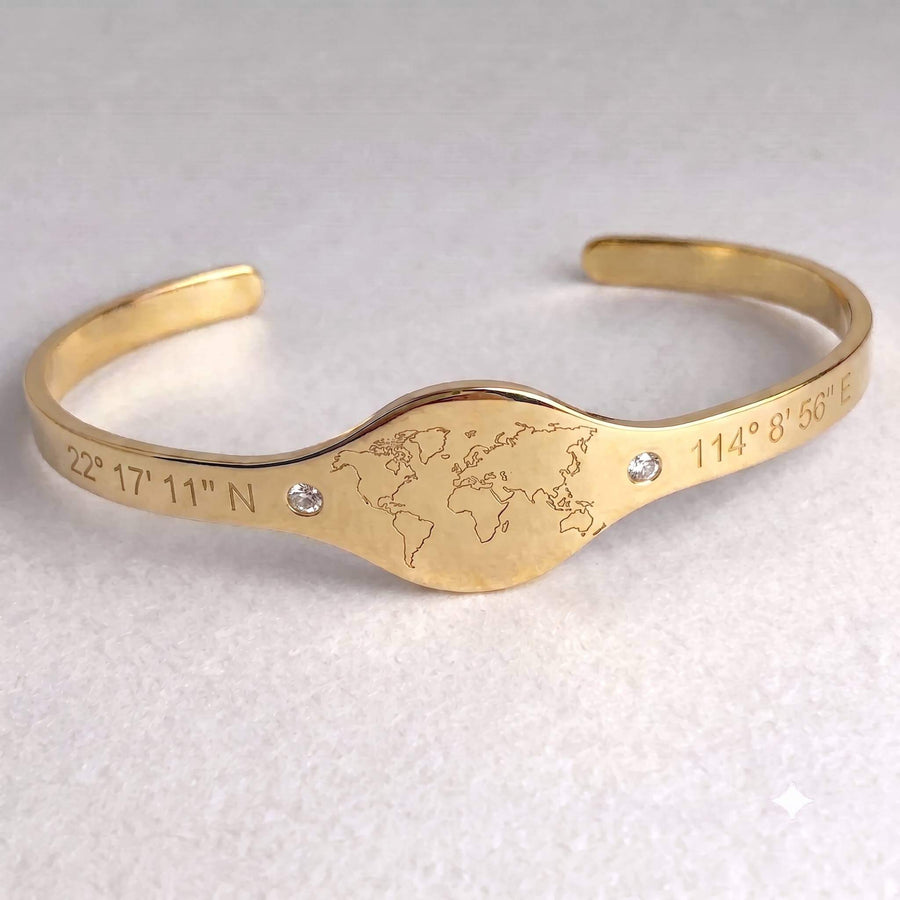 A gold-colored open cuff bracelet featuring an engraved world map in the center, flanked by engraved geographic coordinates, with two small clear rhinestones on a light textured background.