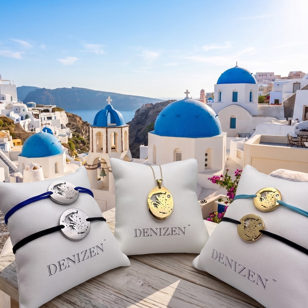 Three white pillows in front of a Santorini coastal scene, each holding a DENIZEN bracelet or pendant. The center pillow shows a gold-toned oval pendant on a chain, while the side pillows show bracelets with round charms in silver and gold tones, plus blue, black, and teal cords.