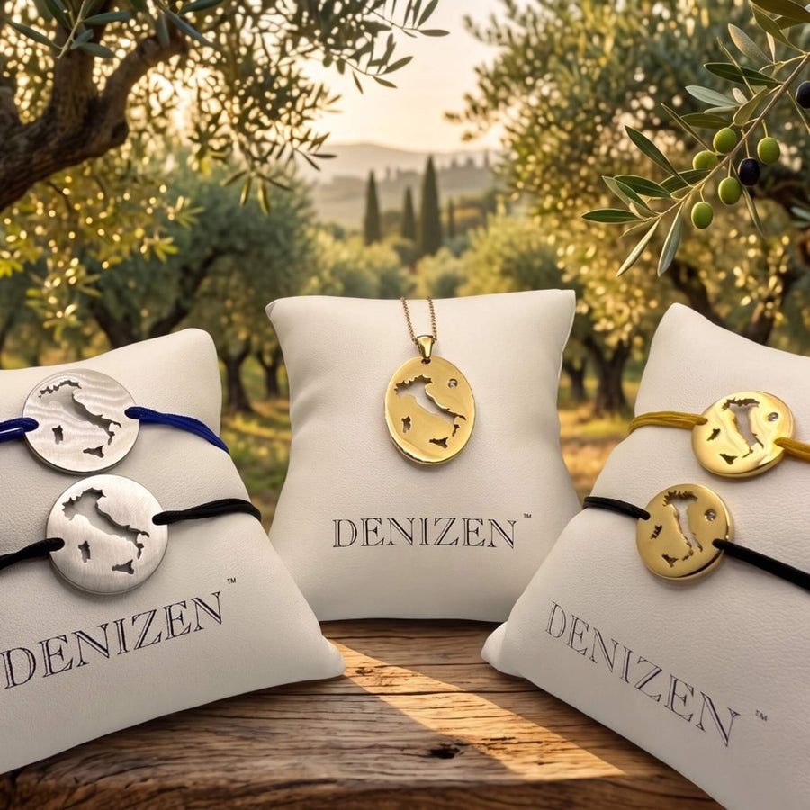Denizen jewelry collection of Italy with map cutouts of oval pendant necklace and bracelets displayed on white pillows in an outdoor olive grove setting. The center pendant is gold and the side pendants are silver on black, navy and beige strings.
