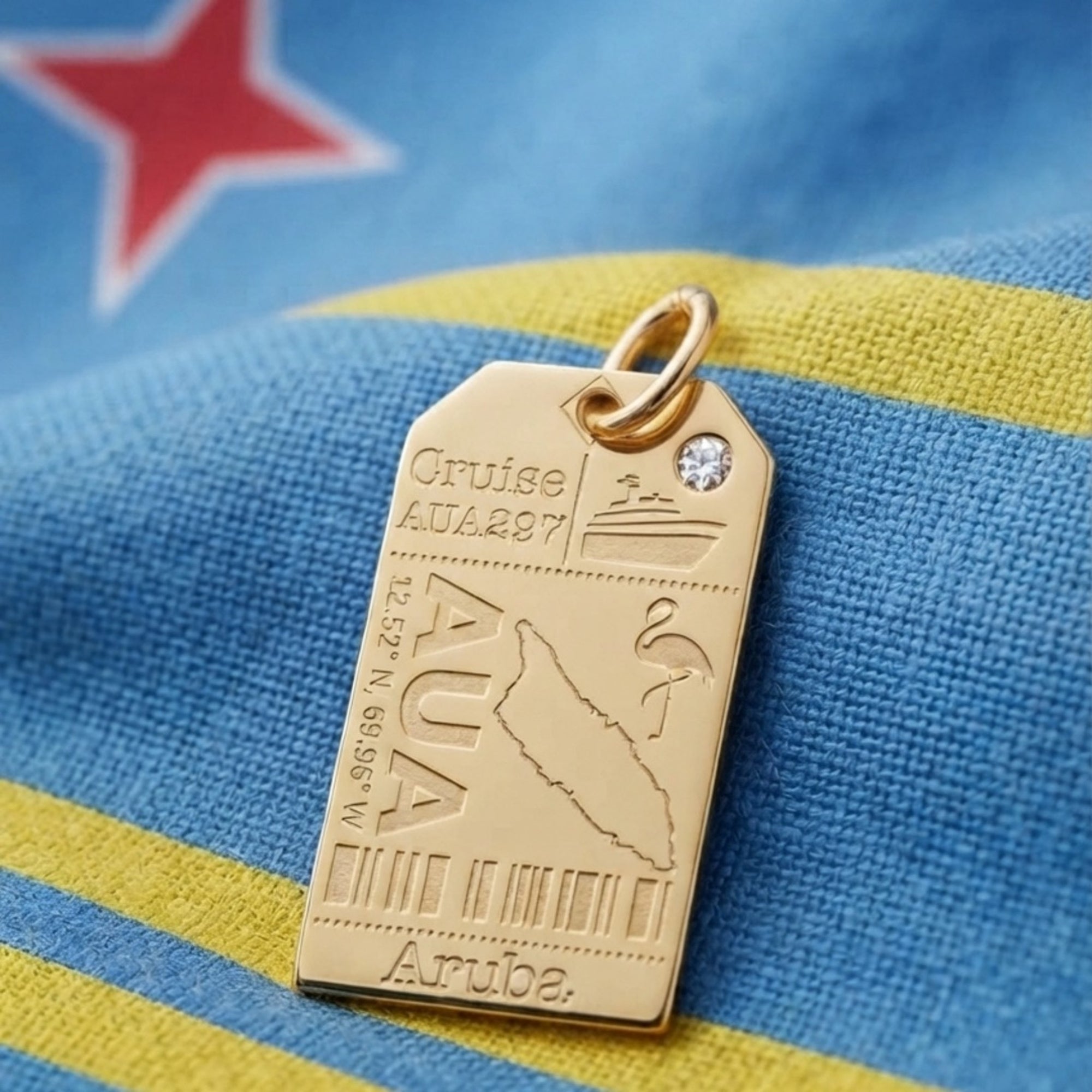 A gold luggage tag charm engraved with with Aruba codes, map and a flamingo and set with a small clear stone.