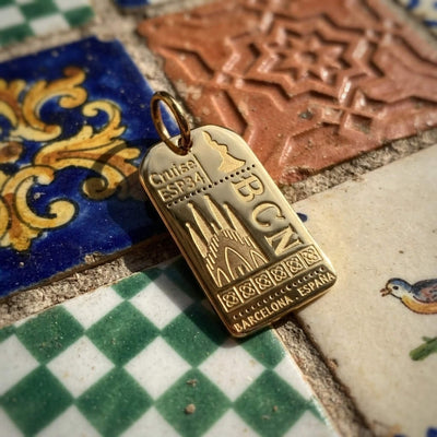 A gold-toned Spain travel souvenir pendant charm shaped like a luggage tag, featuring the text 'BARCELONA - ESPAÑA' and engraved Sagrada Familia and flamenco dancer, resting on a colorful tiled surface.
