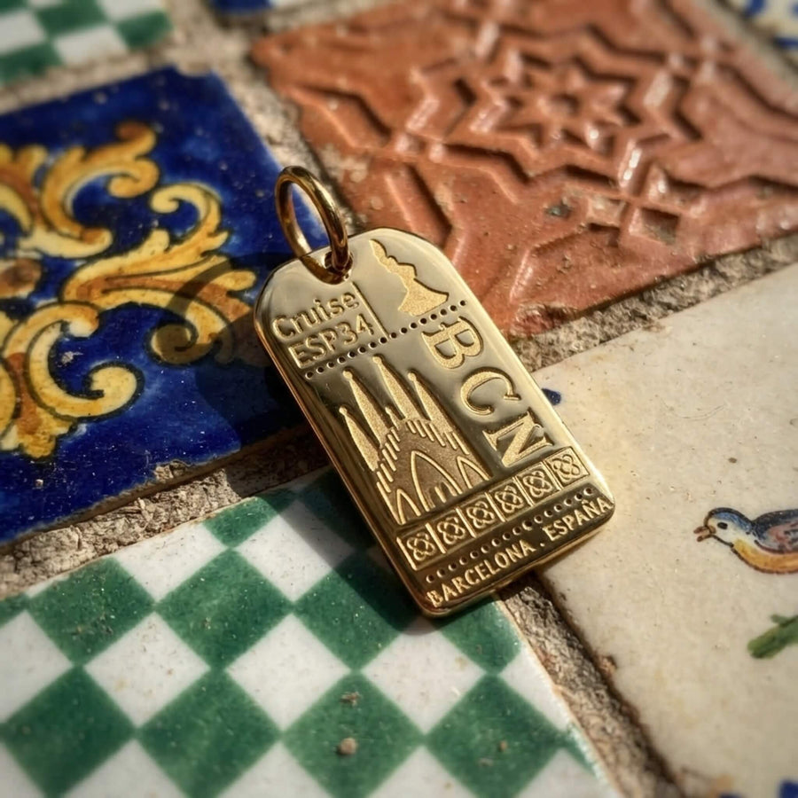 A silver signpost charm and a gold postcard charm displayed outdoors in front of Sagrada Familia in Barcelona. 
