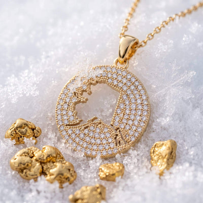 A round gold pendant necklace pavé-set design, displayed on snow with scattered gold nuggets. The pendant features a central cutout of the Alaska map.