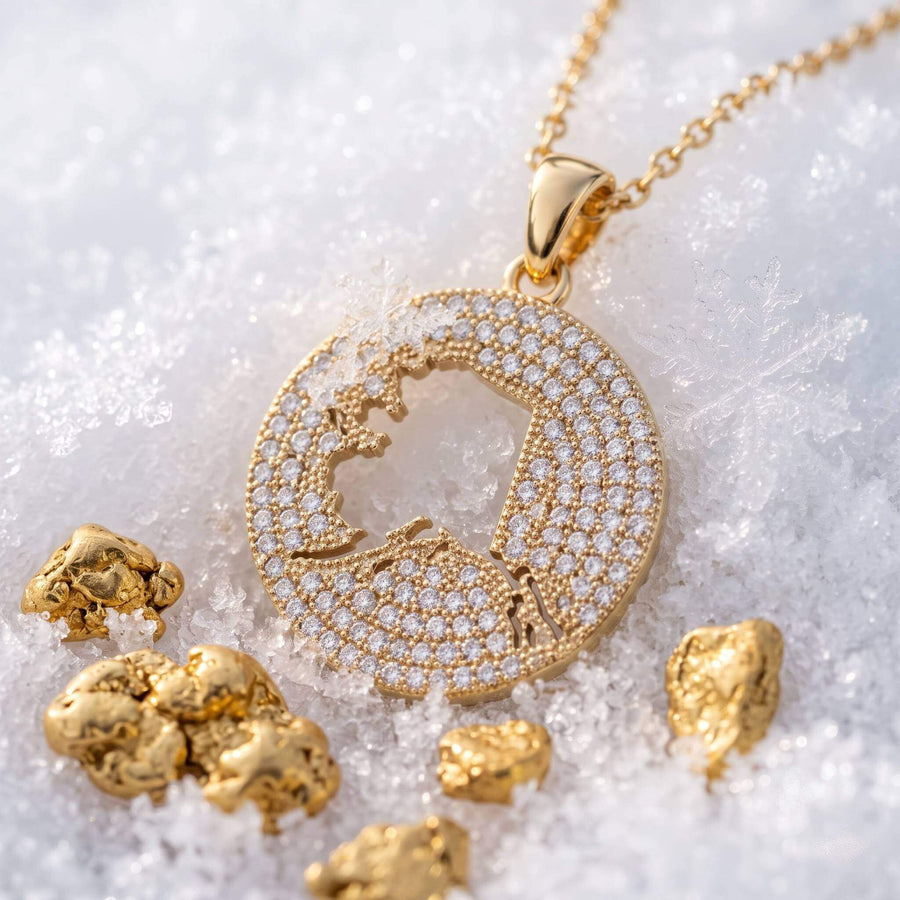 A round gold pendant necklace pavé-set design, displayed on snow with scattered gold nuggets. The pendant features a central cutout of the Alaska map.