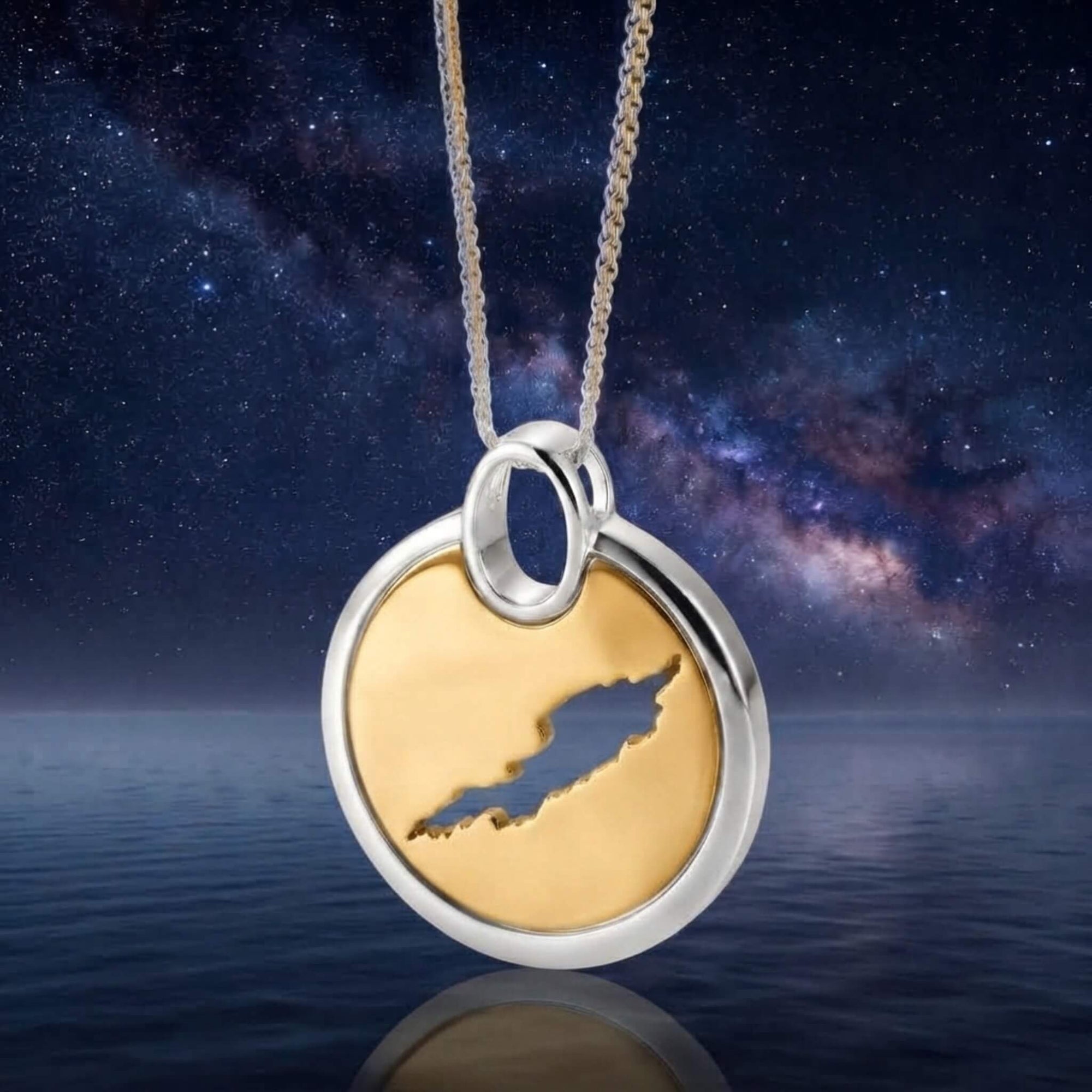 A round gold pendant necklace featuring a cut-out map of Anguilla inside a silver circular frame, suspended from a silver chain against a starry night sky and water background.