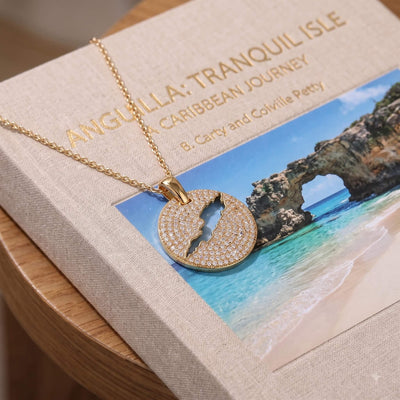 A gold necklace with a round pavé pendant featuring a cut-out map of Anguilla island, displayed on a textured beige book cover with a tropical beach photo.