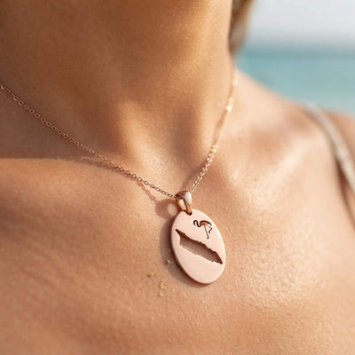 A close-up lifestyle shot of a woman's neck and chest at the beach, wearing a rose gold oval pendant necklace with a cutout design of a flamingo. and the map of Aruba