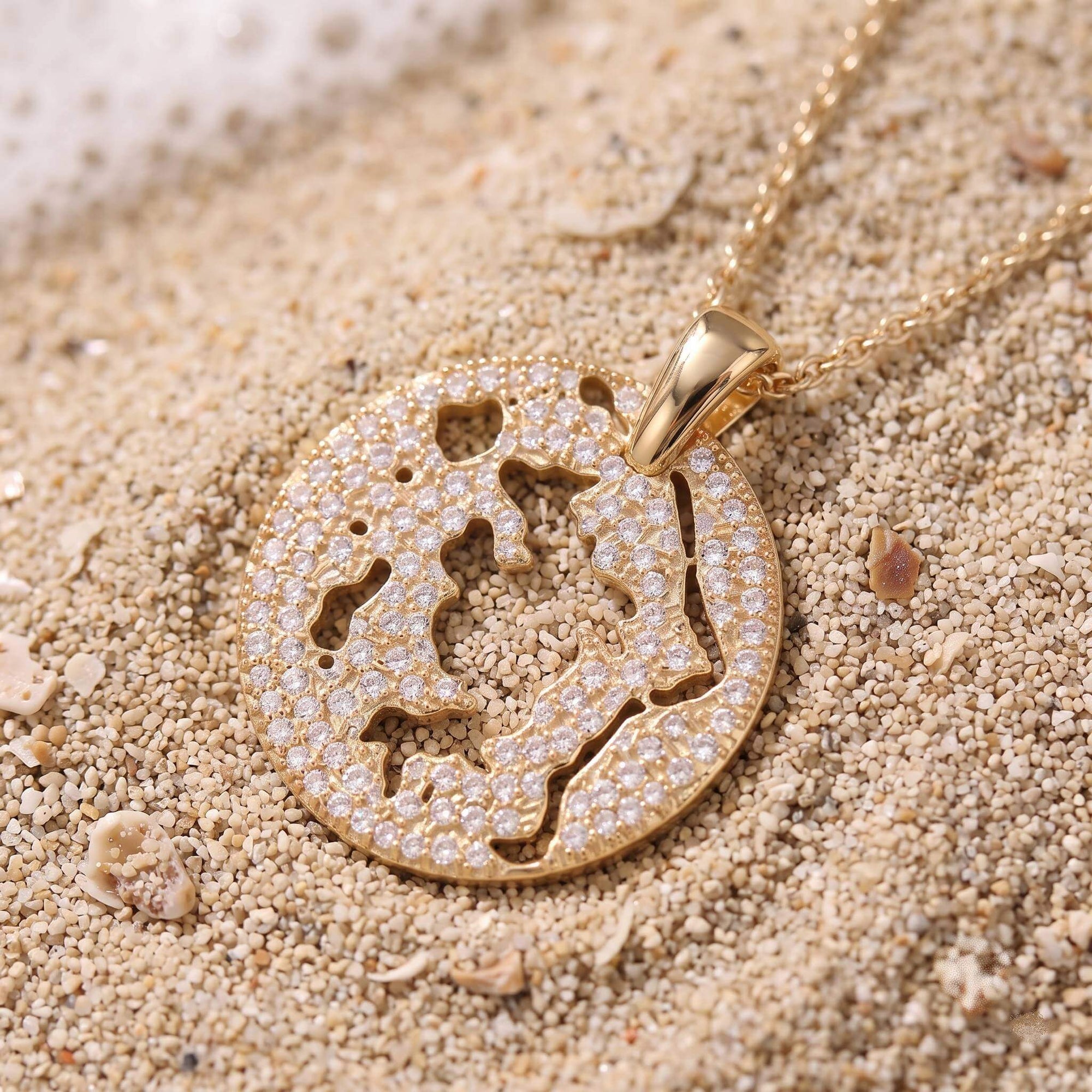 A gold pendant necklace with a cutout map of Bora Bora in a diamond pavé, resting on sandy beach ground with small shells.