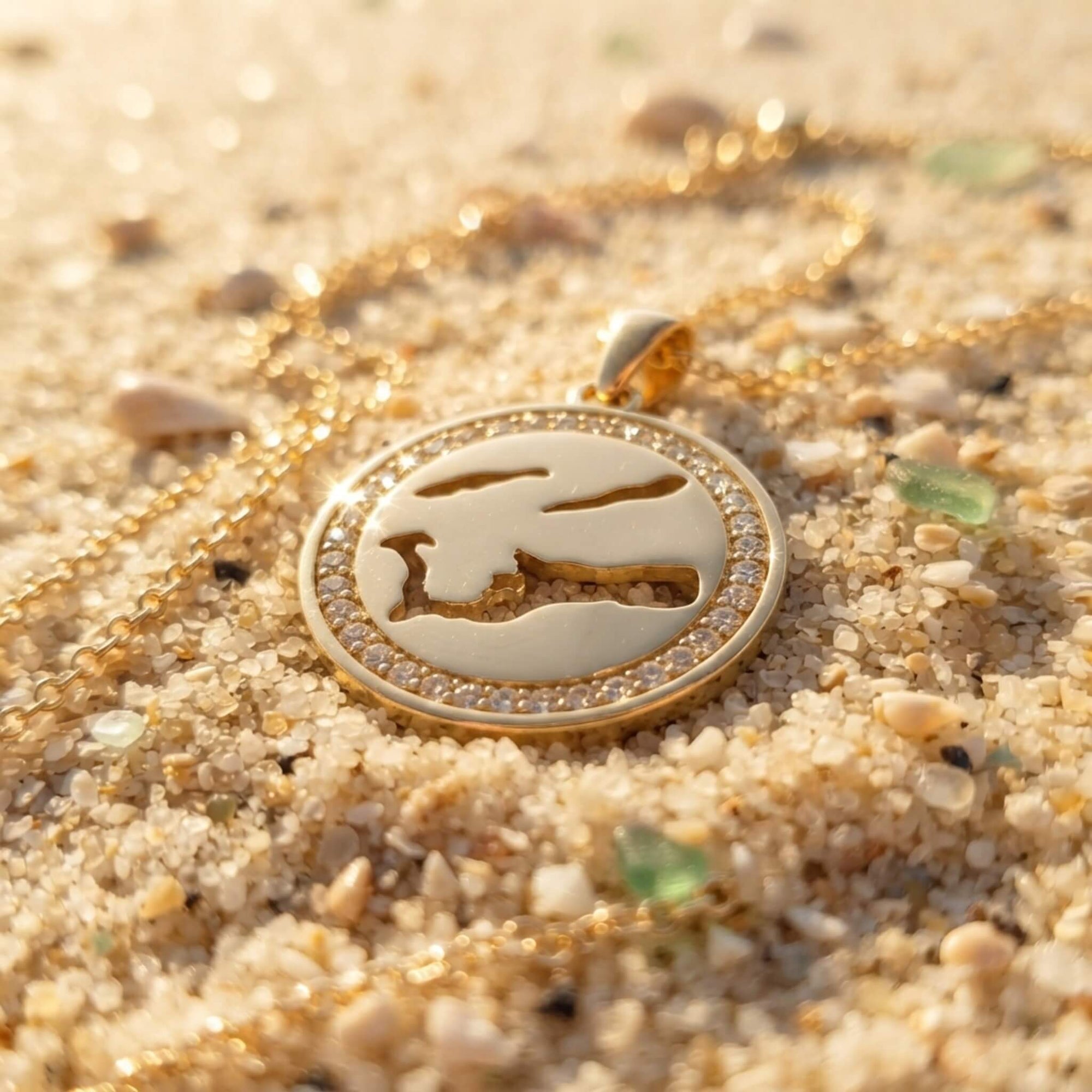 A gold round pendant necklace with a cut-out map of Cayman islands and an outer ring of clear stones resting on sand.