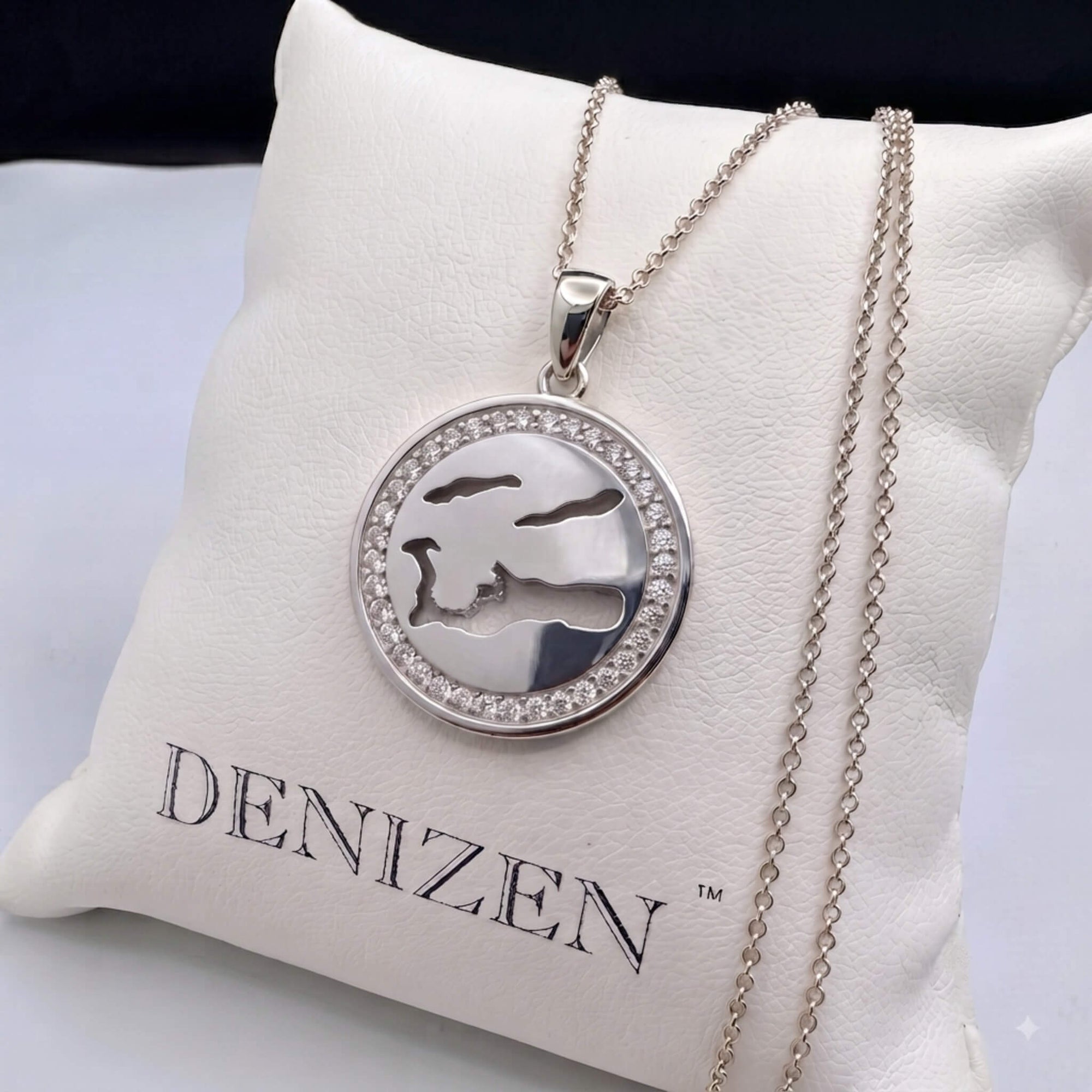 A silver round pendant necklace with a cutout design of the Cayman Islands map and a border of small clear stones, displayed on a white cushion printed with the brand name DENIZEN.