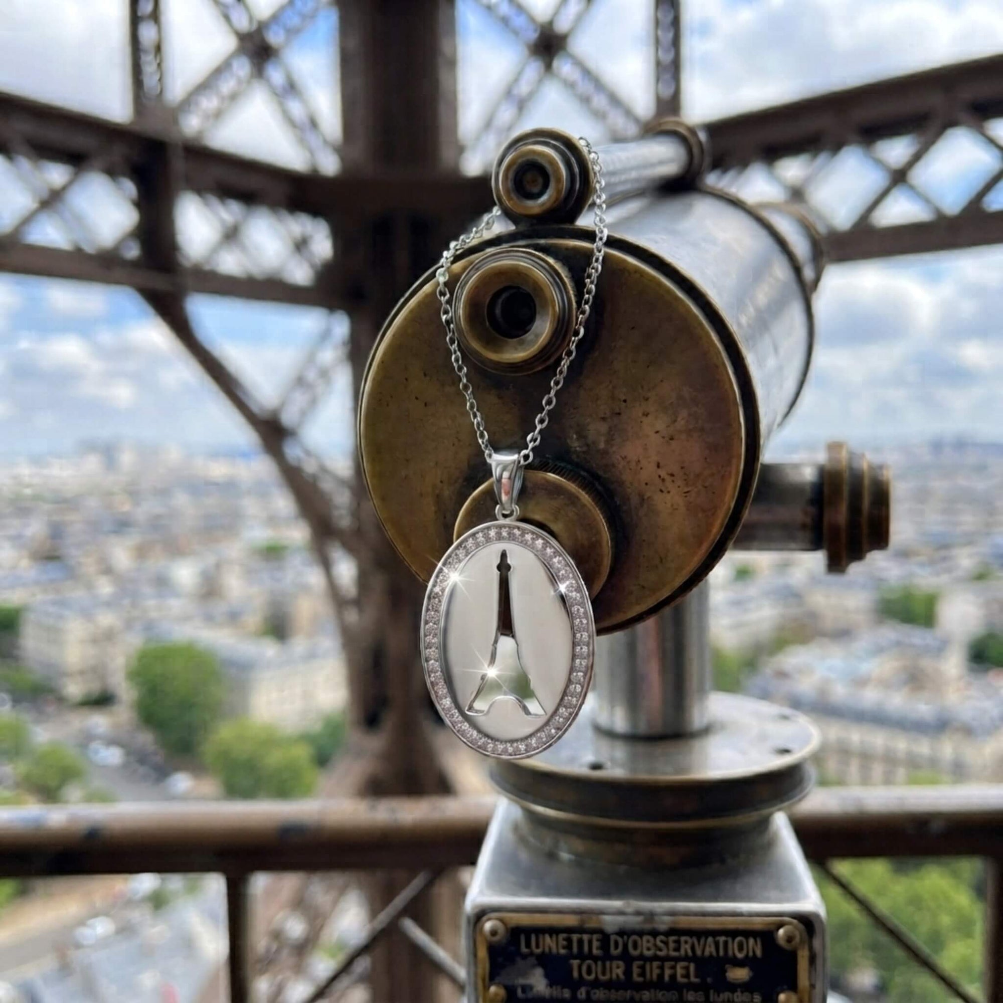 A silver oval pendant necklace with a clear crystal border and Eiffel Tower cutout, draped over a brass telescope at the Eiffel Tower with a Paris cityscape in the background.