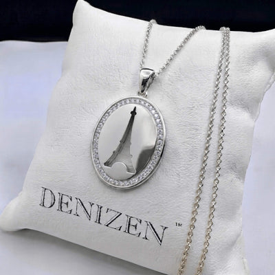 A silver oval pendant necklace with a cutout Eiffel Tower design and a border of small clear stones, displayed on a white cushion printed with the brand name DENIZEN.