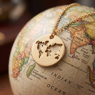 A gold round pendant necklace with a cutout world map design, resting on a vintage-style globe map.