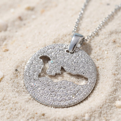 A silver pavé round pendant necklace with a cut-out map of Grand Cayman island, resting on light beige sand.