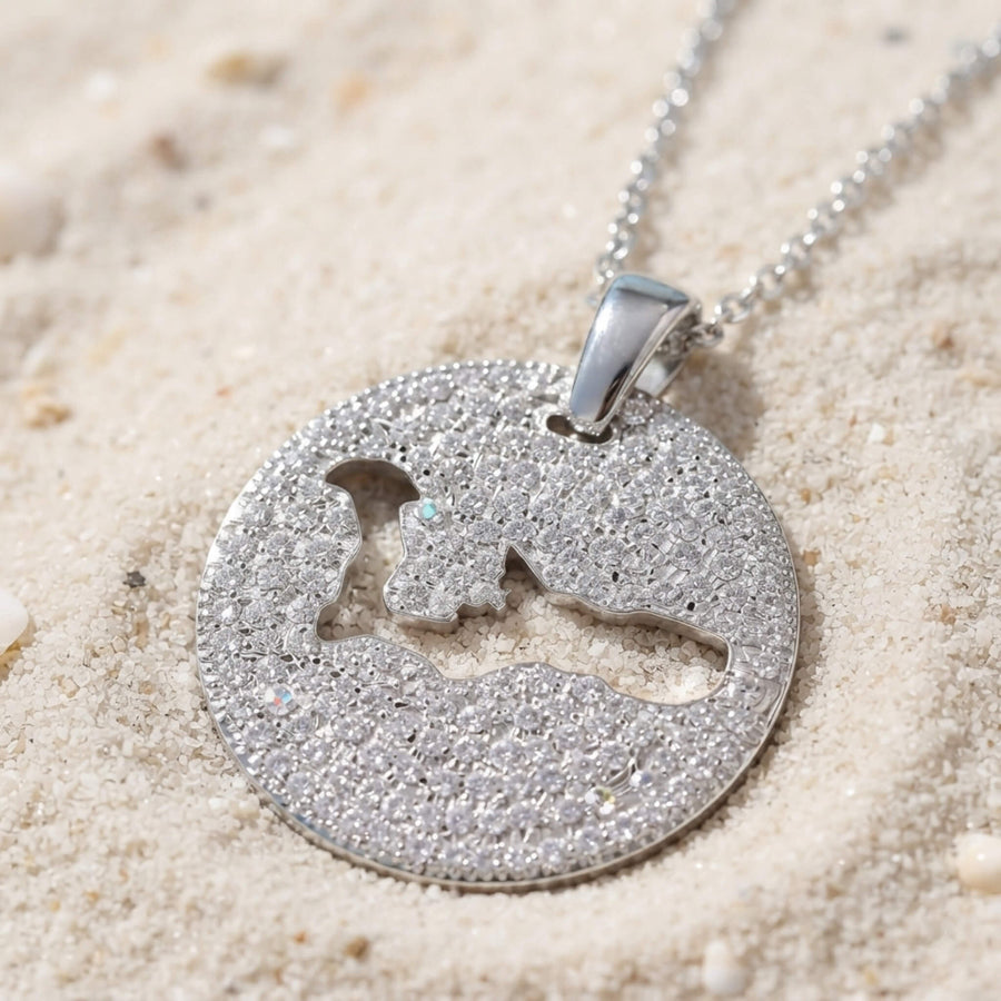 A silver pavé round pendant necklace with a cut-out map of Grand Cayman island, resting on light beige sand.