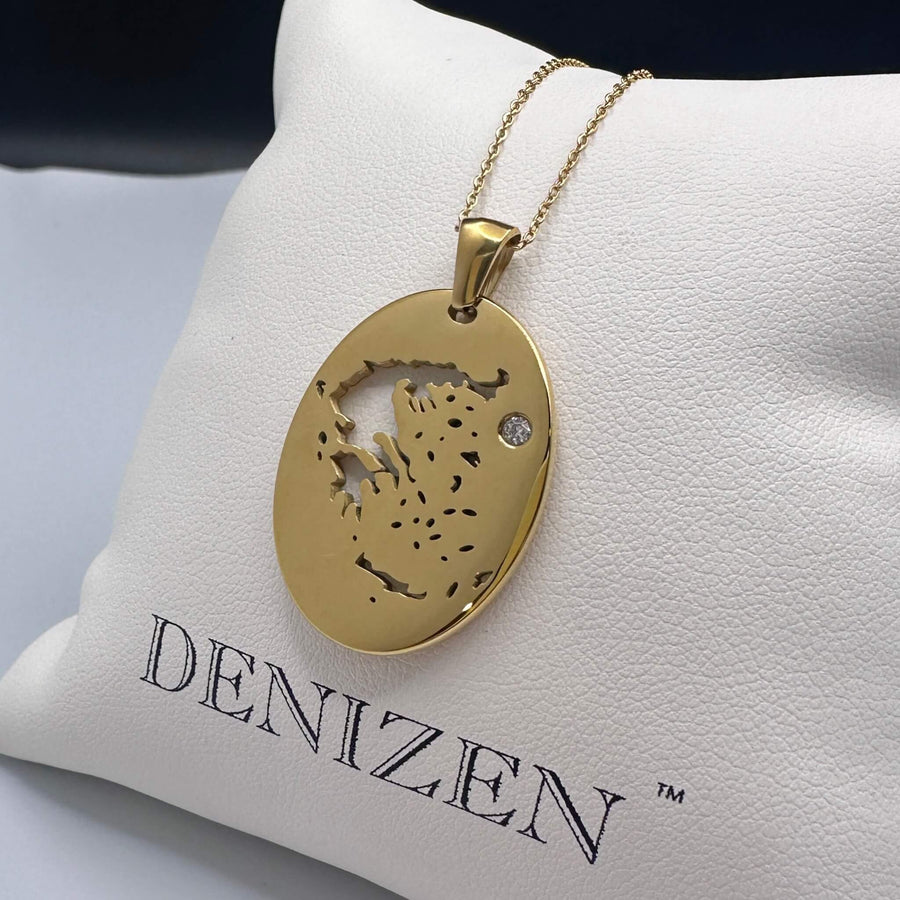 Three white pillows in front of a Santorini coastal scene, each holding a DENIZEN bracelet or pendant. The center pillow shows a gold-toned oval pendant on a chain, while the side pillows show bracelets with round charms in silver and gold tones, plus blue, black, and teal cords.