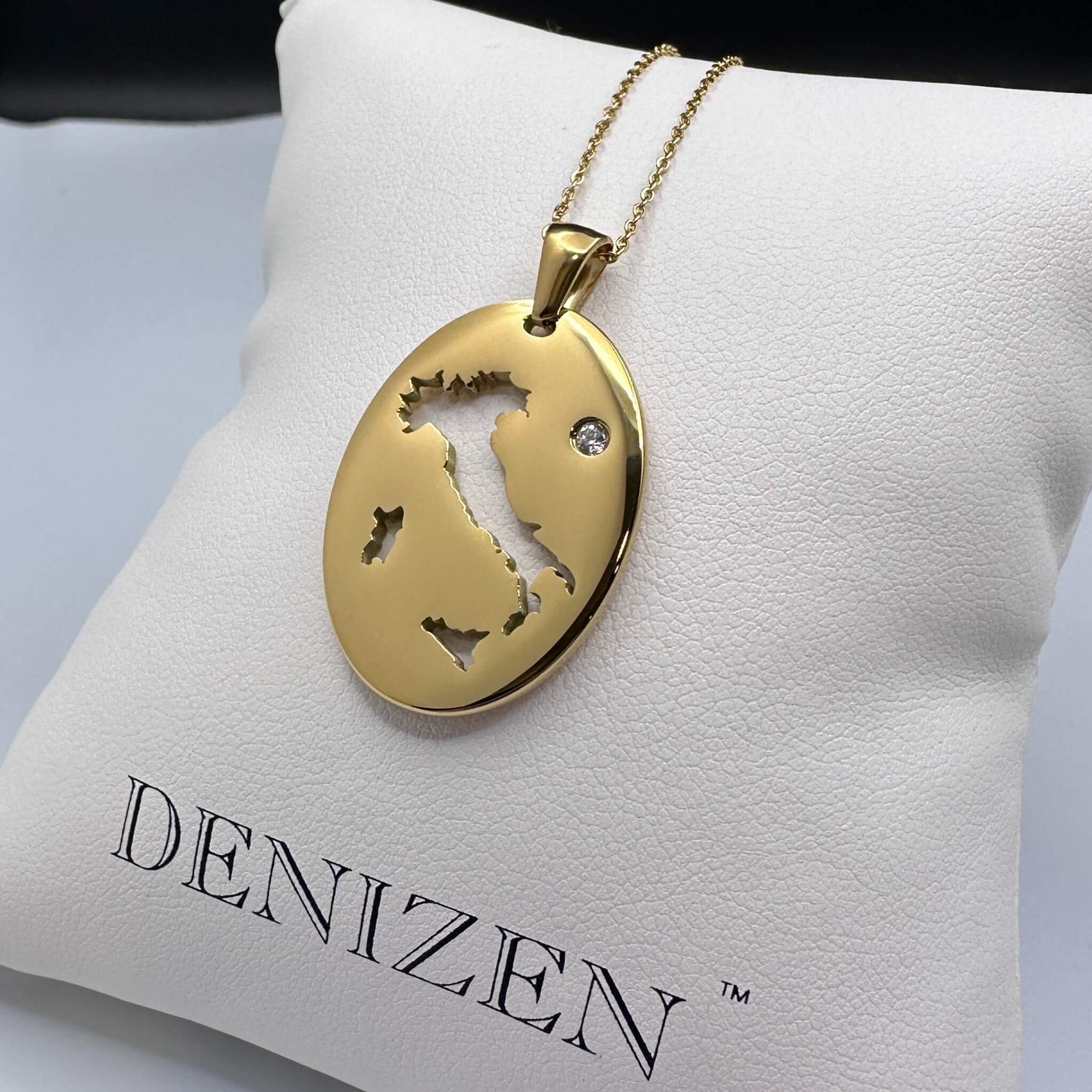 A gold oval pendant necklace with a cut-out map of Italy and a small clear stone, displayed on a white Denizen jewelry cushion.
