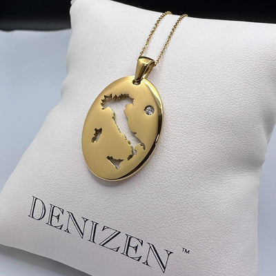 A gold oval pendant necklace with a cut-out map of Italy and a small clear stone, displayed on a white Denizen jewelry cushion.