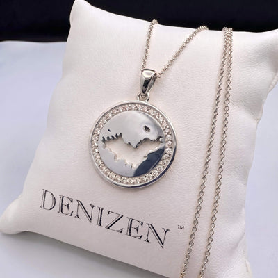 A silver pendant necklace with a St-Barth map cutout design and an outer ring of small clear stones, displayed on a white cushion printed with the brand name DENIZEN.
