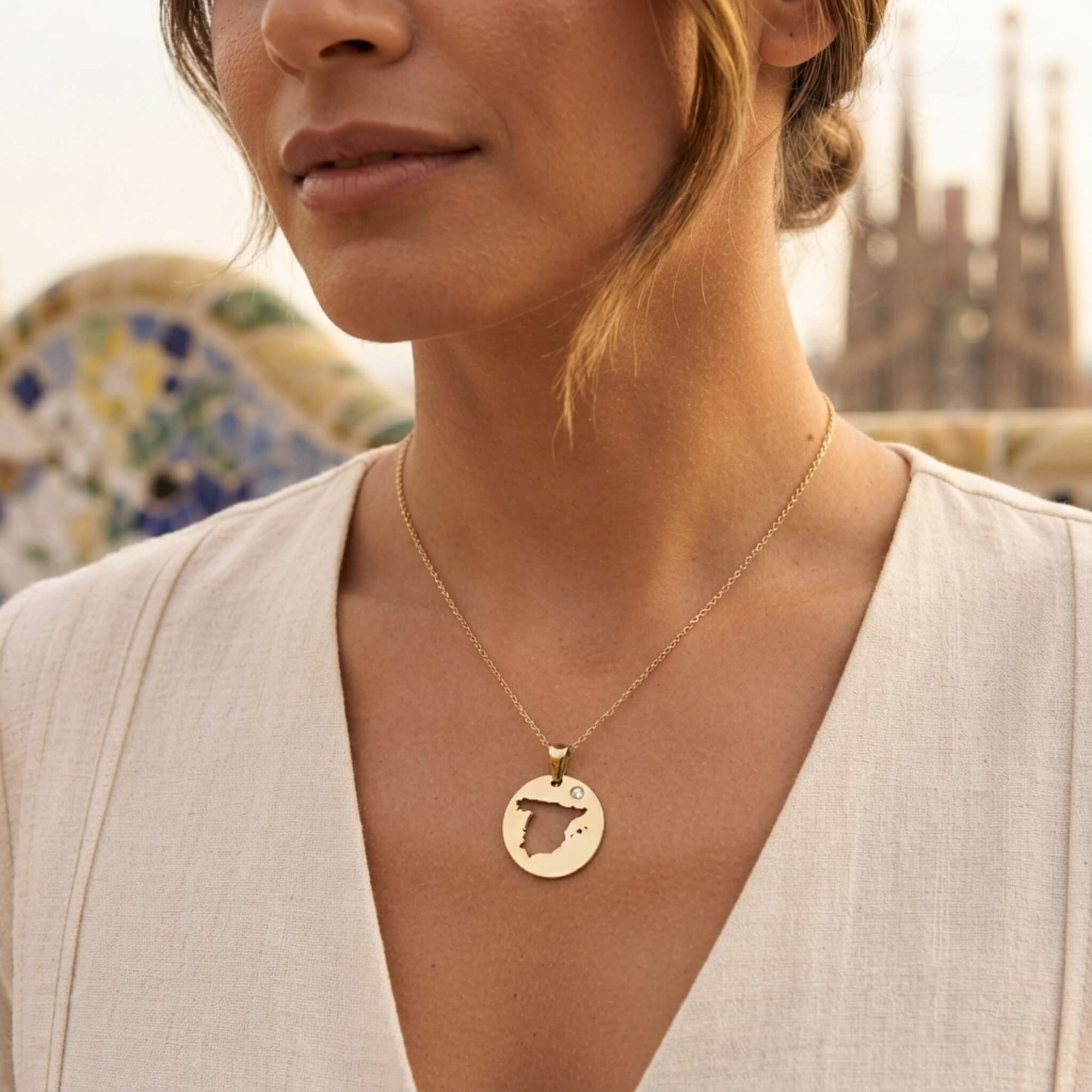 A close-up lifestyle shot of a woman wearing a delicate gold chain necklace with a circular pendant featuring a cut-out map of Spain, set against a softly blurred outdoor background. of Barcelona