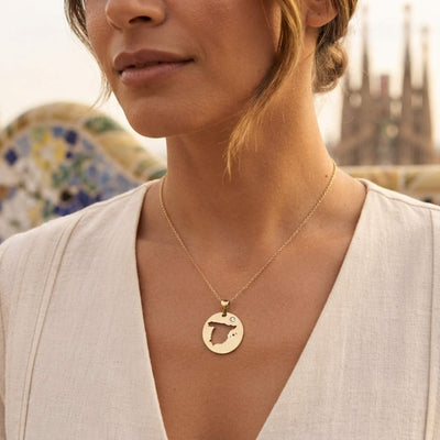 A close-up lifestyle shot of a woman wearing a delicate gold chain necklace with a circular pendant featuring a cut-out map of Spain, set against a softly blurred outdoor background. of Barcelona