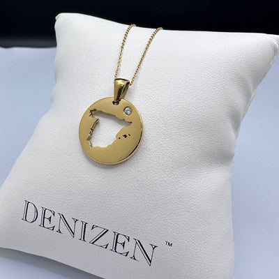 A Denizen gold chain pendant with a cut-out map of Spain displayed on a white branded cushion.