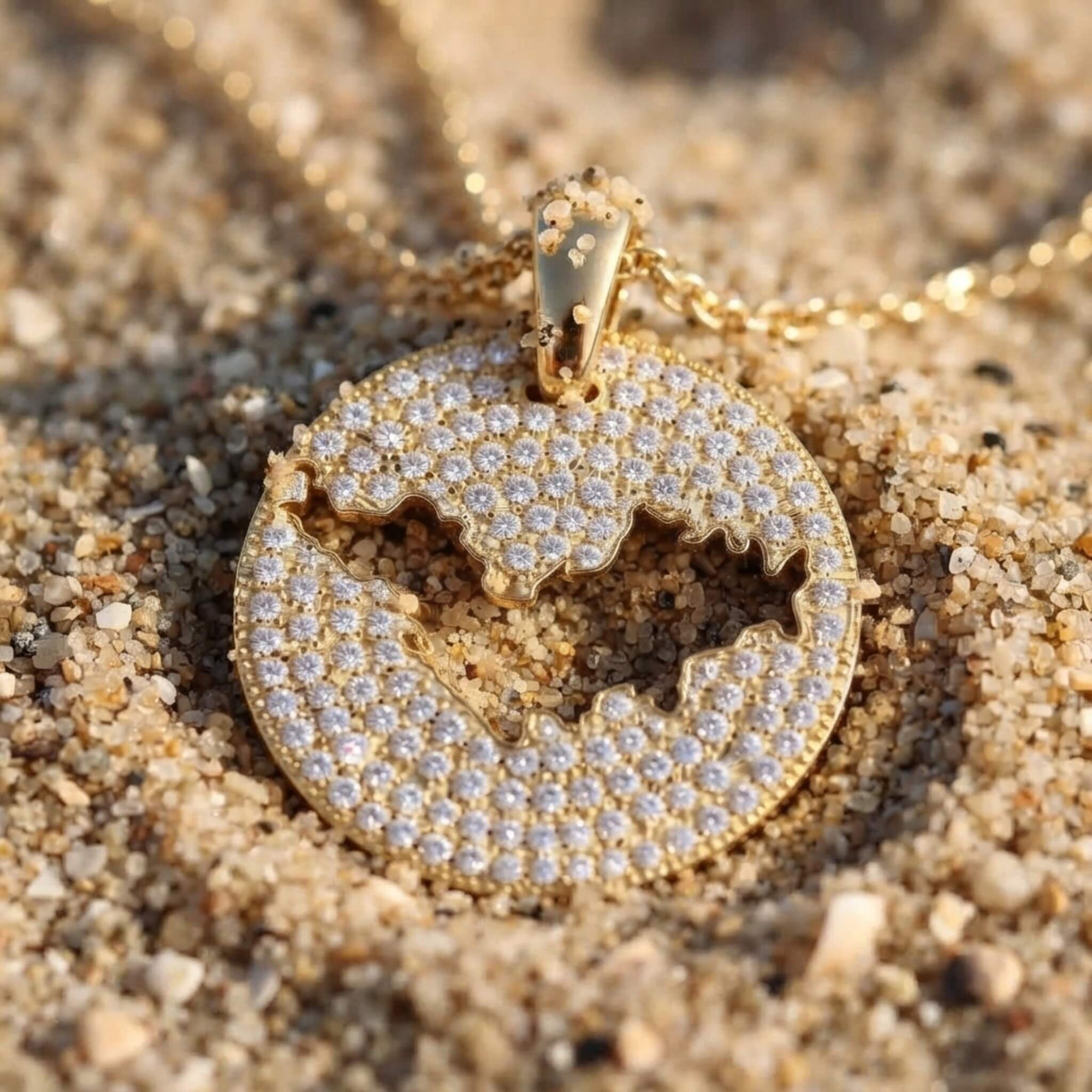 A round gold pendant necklace covered in clear pavé stones, featuring a cutout of St-Barth map, resting on sand.