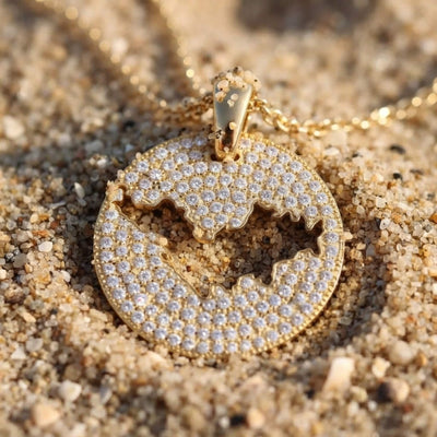 A round gold pendant necklace covered in clear pavé stones, featuring a cutout of St-Barth map, resting on sand.
