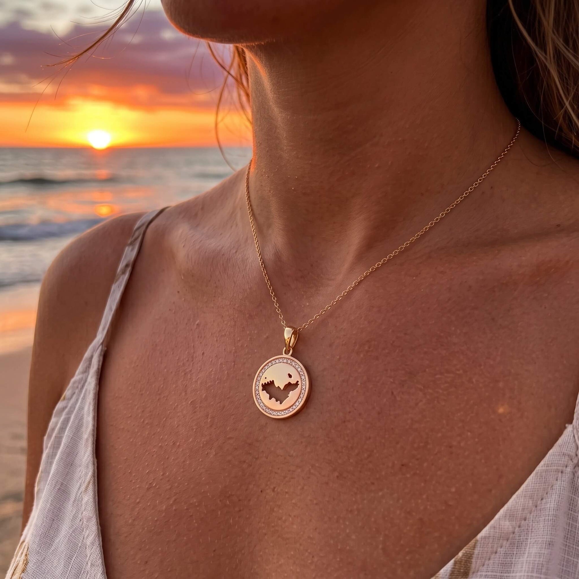 A gold-toned necklace with a circular pendant featuring a cutout design of the map of St-Barth, worn by a woman at sunset by the beach.