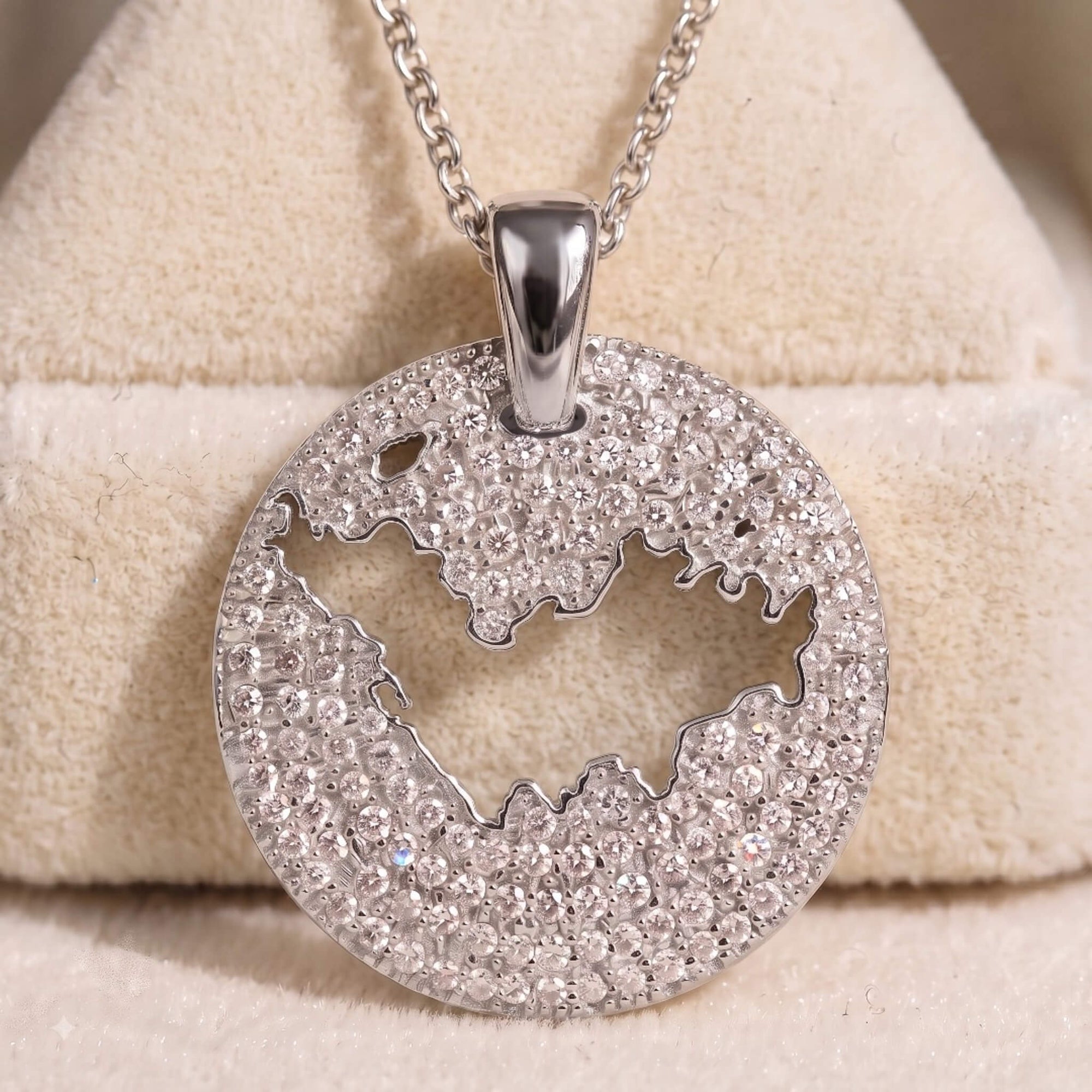 A round silver pendant necklace covered in clear pavé stones, featuring a cutout of St-Barth map, displayed on a beige velvet jewelry stand.
