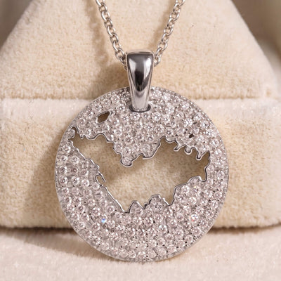 A round silver pendant necklace covered in clear pavé stones, featuring a cutout of St-Barth map, displayed on a beige velvet jewelry stand.