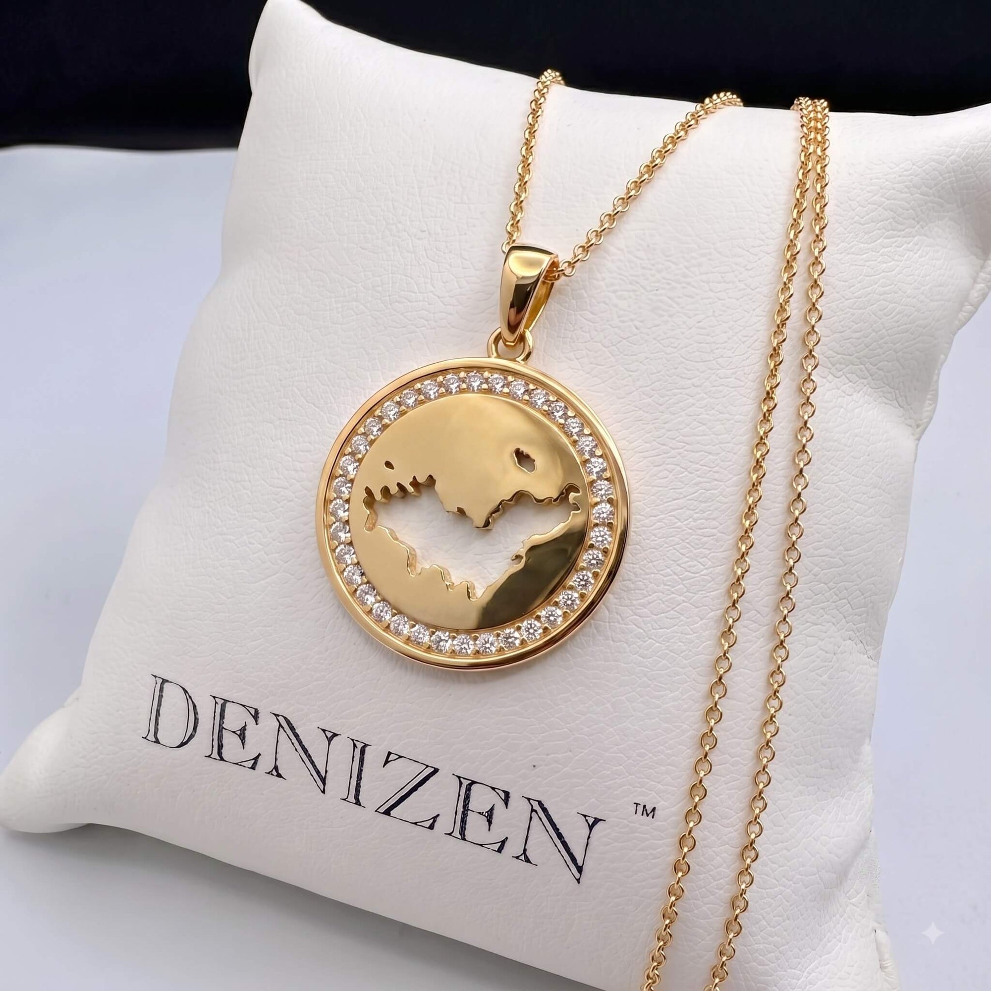 A gold pendant necklace with a St-Barth map cutout design and an outer ring of small clear stones, displayed on a white cushion printed with the brand name DENIZEN.