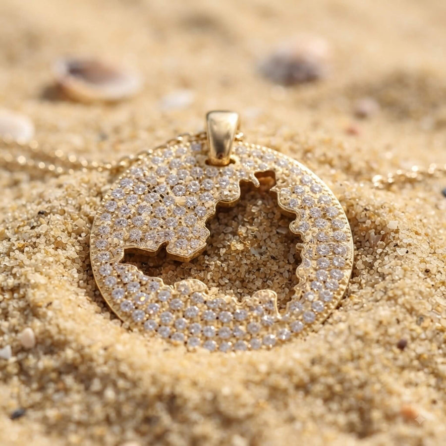 A gold-toned round pendant necklace with a cutout of St. Maarten map, set with small clear stones and resting on sandy beach ground.